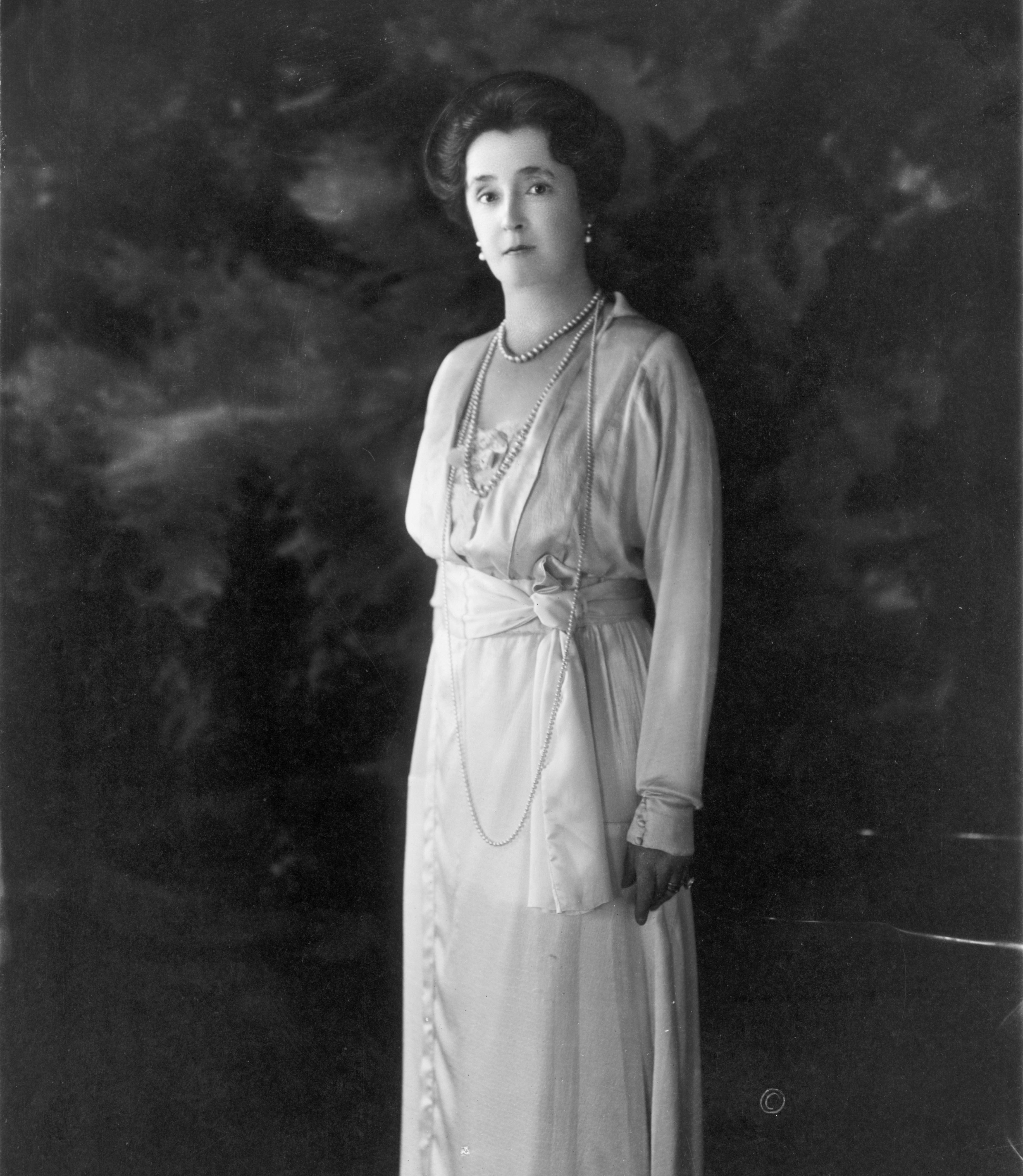 A woman stands elegantly, wearing a light gown with layered necklaces, set against a dark, blurred backdrop.