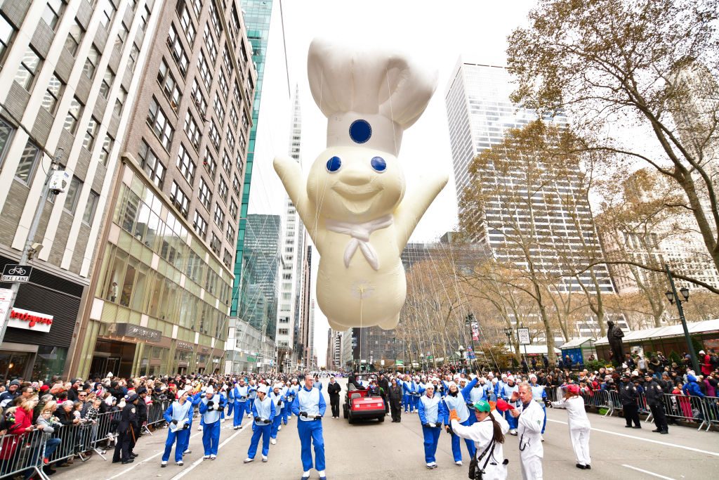 A giant, smiling doughboy balloon floats above a city parade, surrounded by handlers and spectators, amidst tall buildings and bare trees.