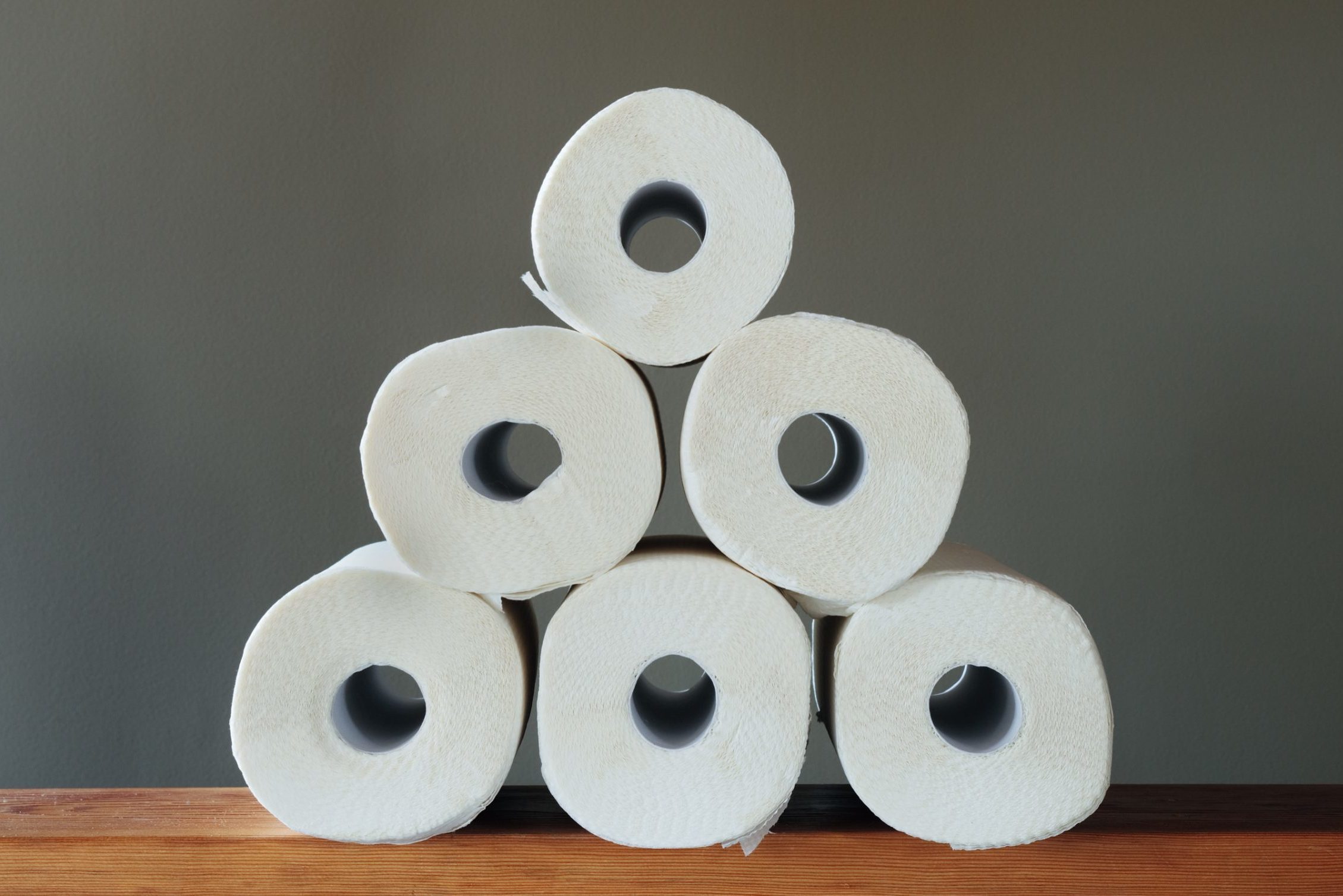 Six toilet paper rolls stacked in a pyramid on a wooden surface against a plain gray background.