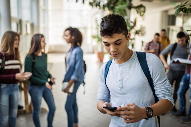 Male high school student using cell phone in a school hallway.