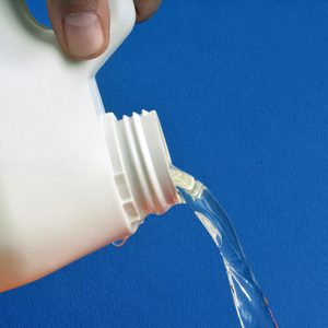 A white container is pouring clear liquid, held by fingertips, against a plain blue background.