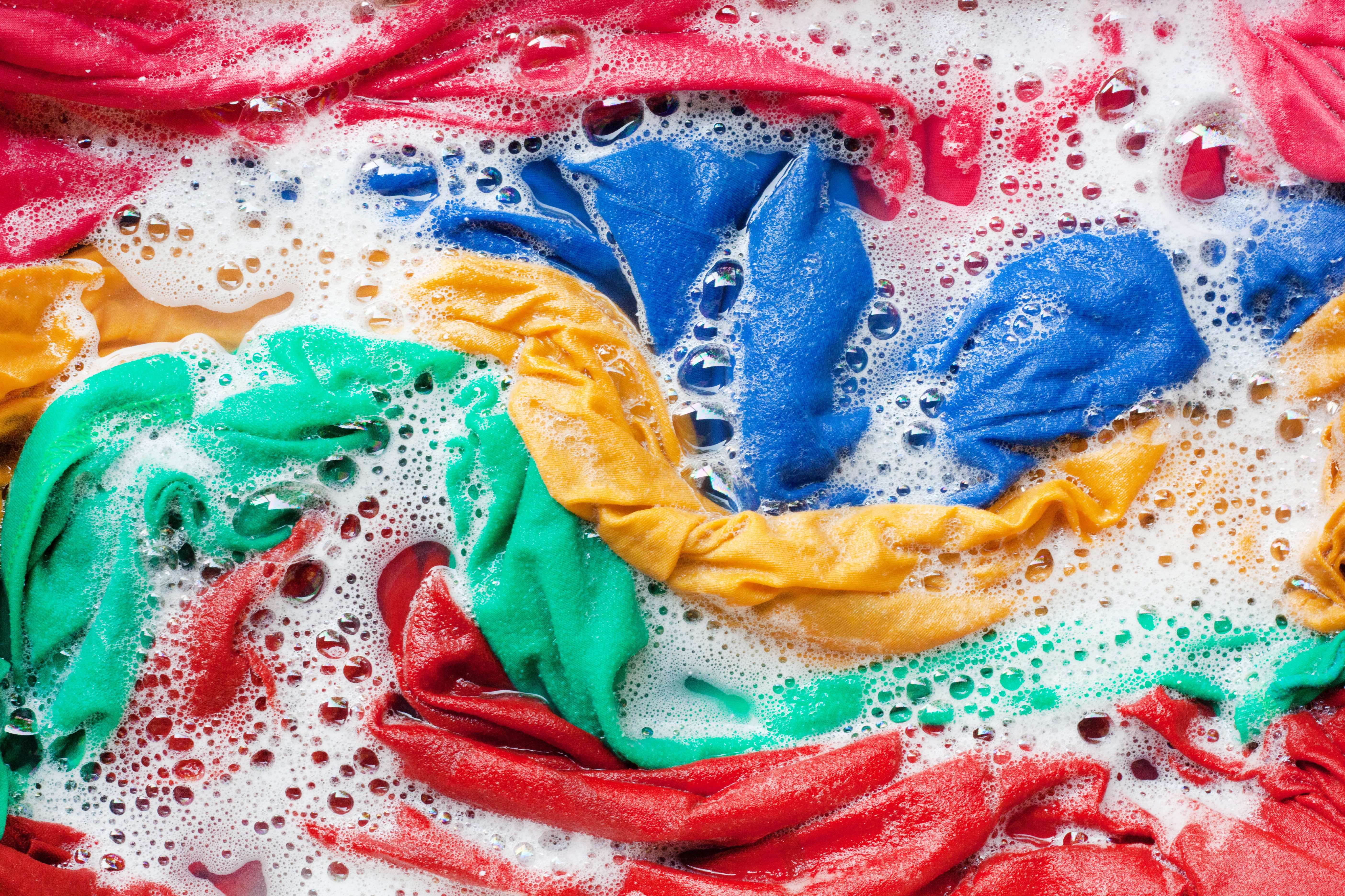 Colorful clothes soak in soapy water with bubbles covering the vibrant fabric; red, blue, yellow, and green cloths are visible.