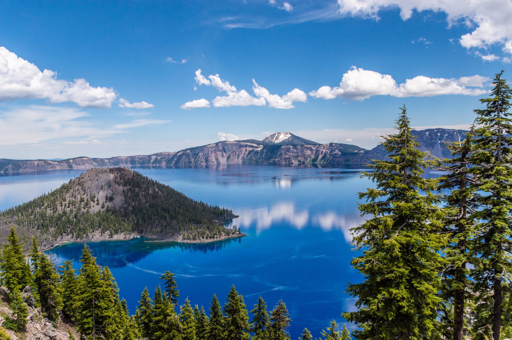 A serene, blue lake reflects distant peaks, surrounded by lush green forests under a clear sky with scattered clouds.