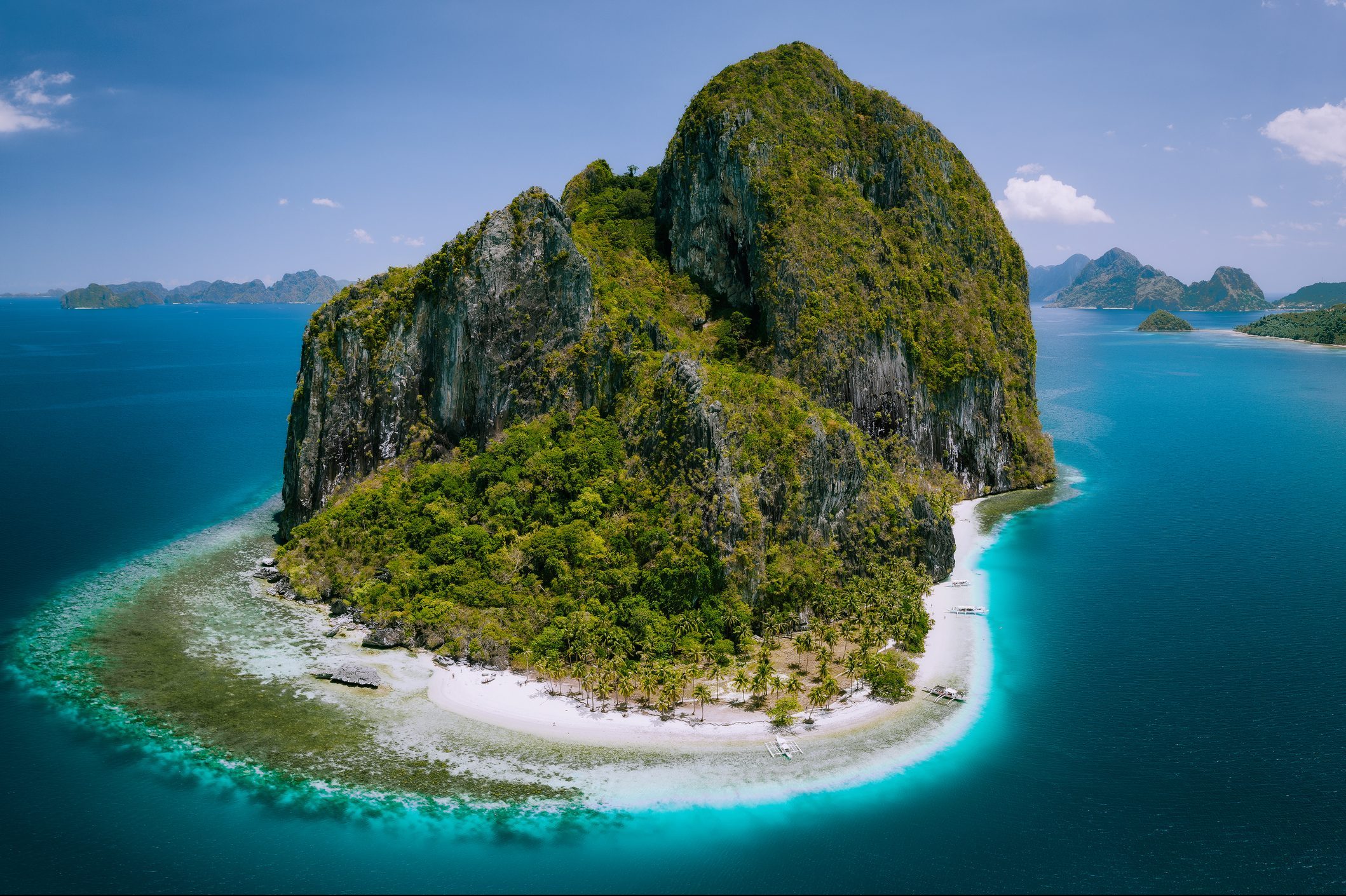 Lush, rocky island surrounded by turquoise sea, featuring white sandy beaches with palm trees, under a clear blue sky with distant islands in view.