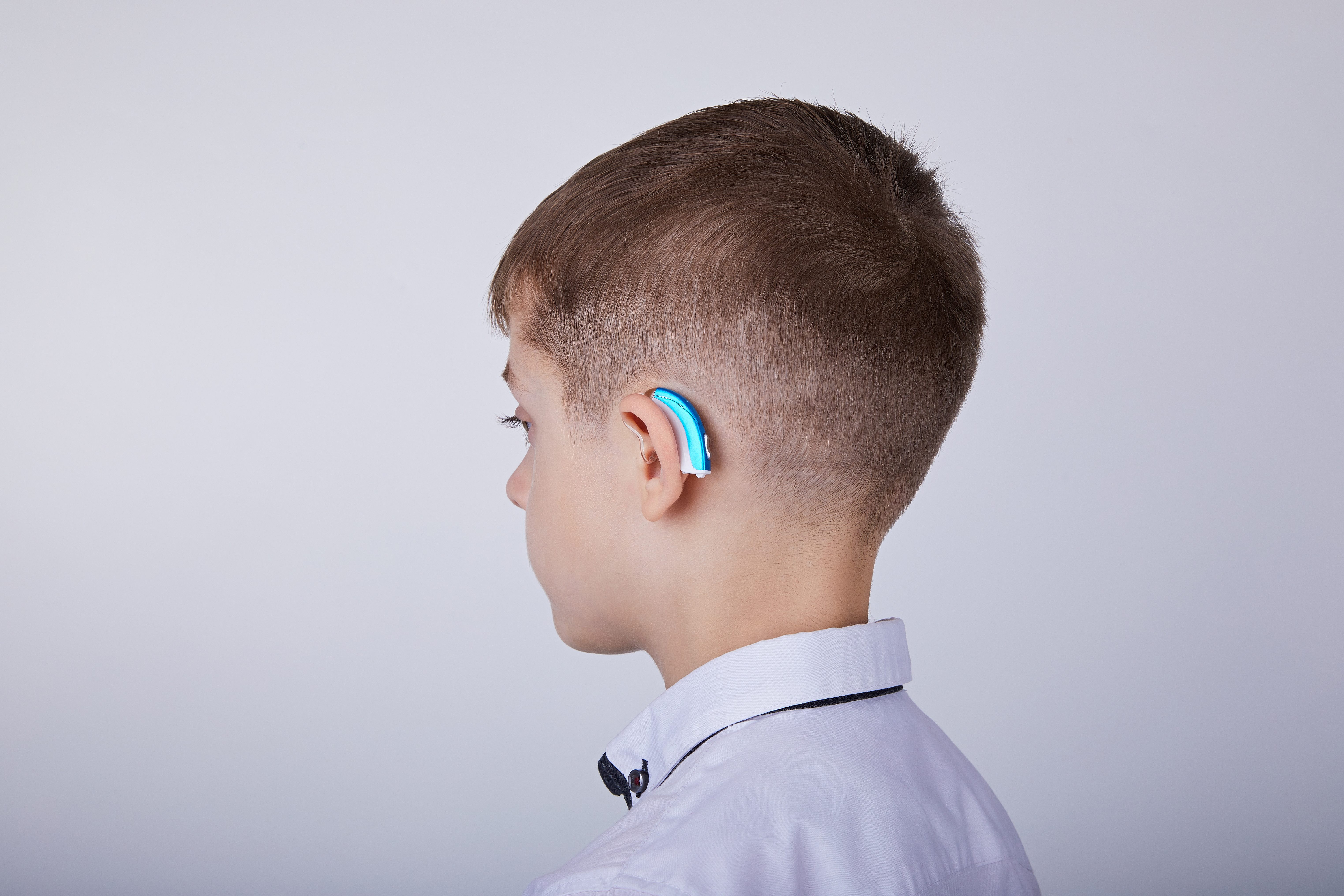 A young boy wearing a blue hearing aid gazes to the side. He is dressed in a white collared shirt against a plain background.