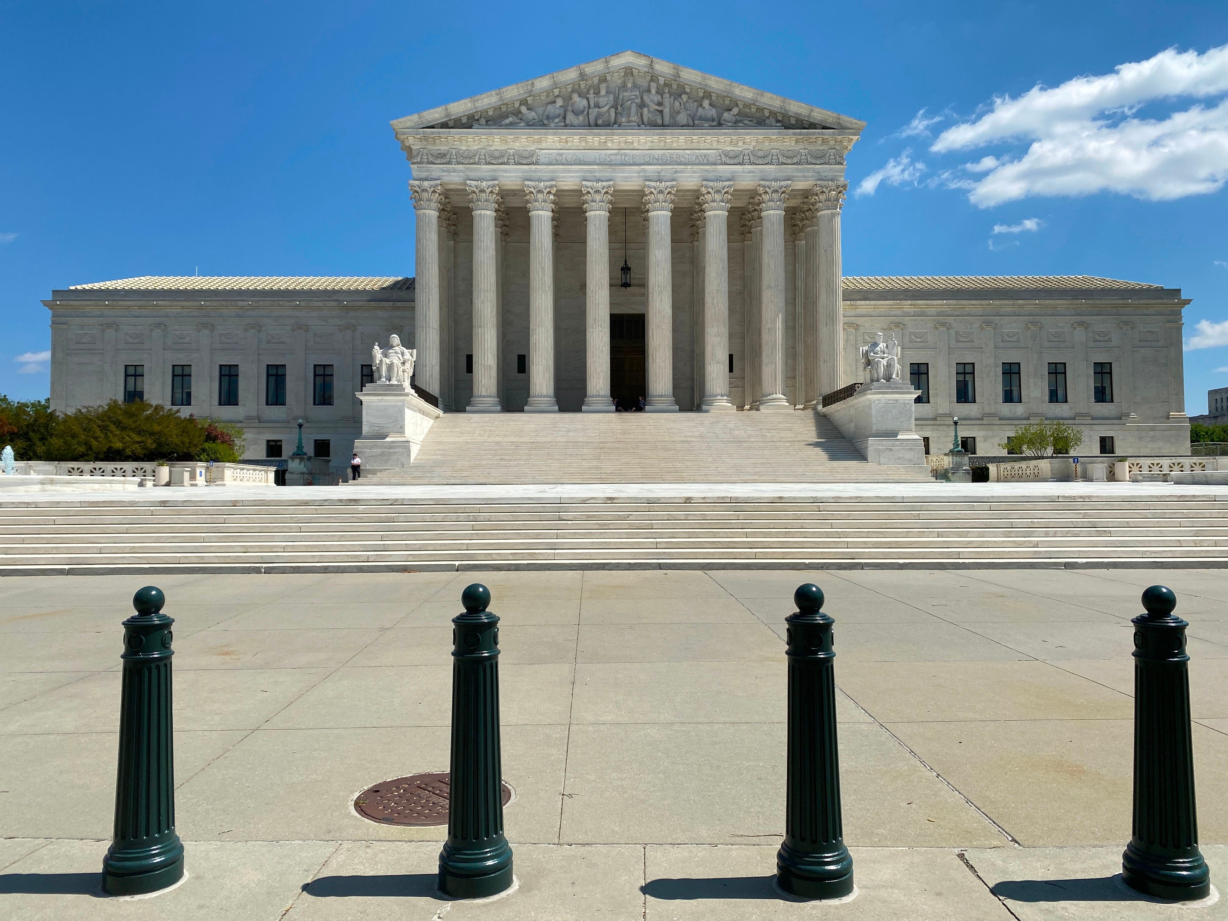 Grand building stands; columns rise; steps lead upward; statues flank entrance; open sky above; paved area and bollards in foreground; serene and stately atmosphere.