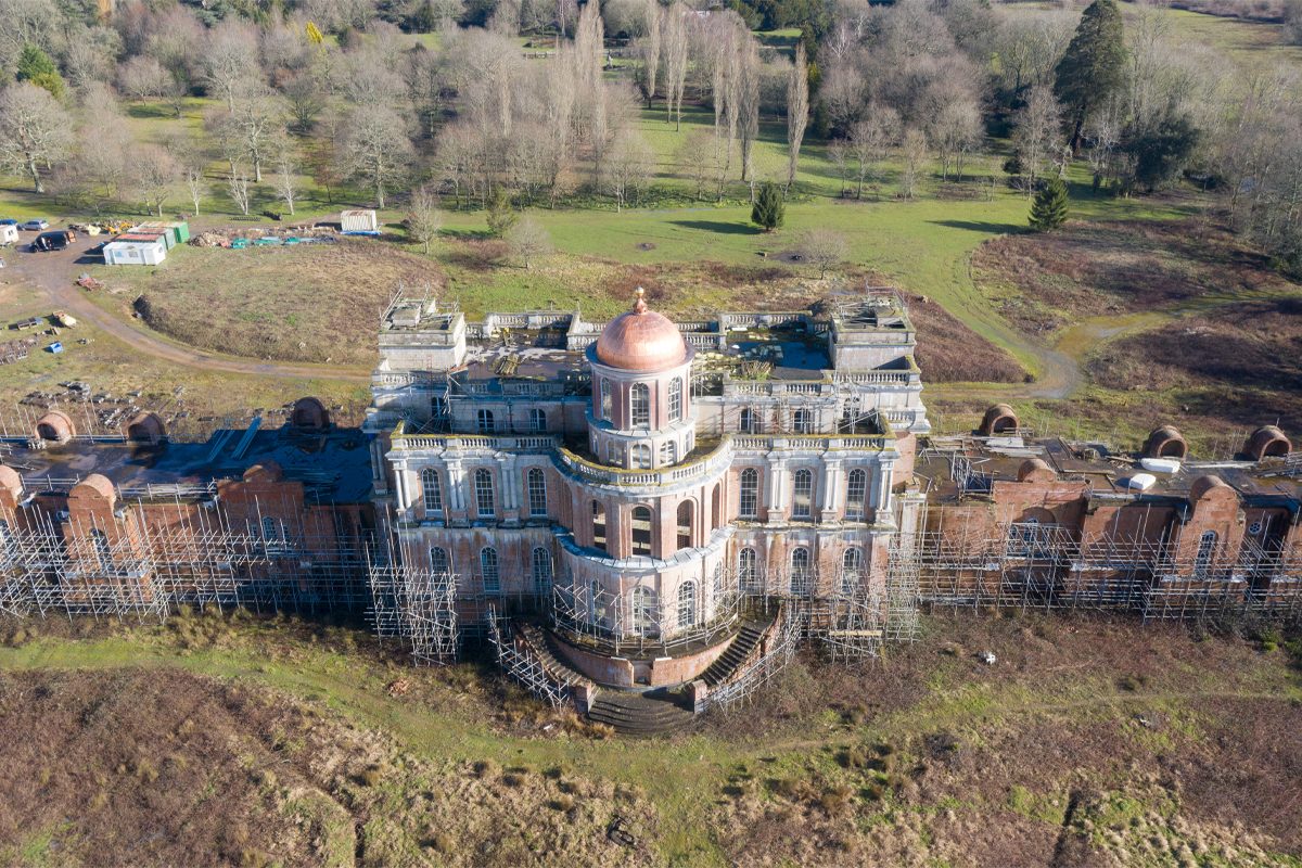Historic building surrounded by scaffolding, showing deterioration, situated in a grassy, wooded landscape.