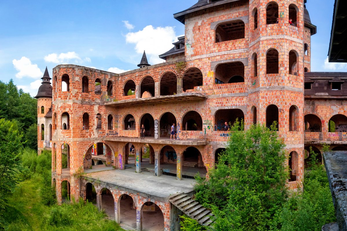 Abandoned castle features arched windows and graffiti-covered walls, surrounded by lush greenery under a bright blue sky.
