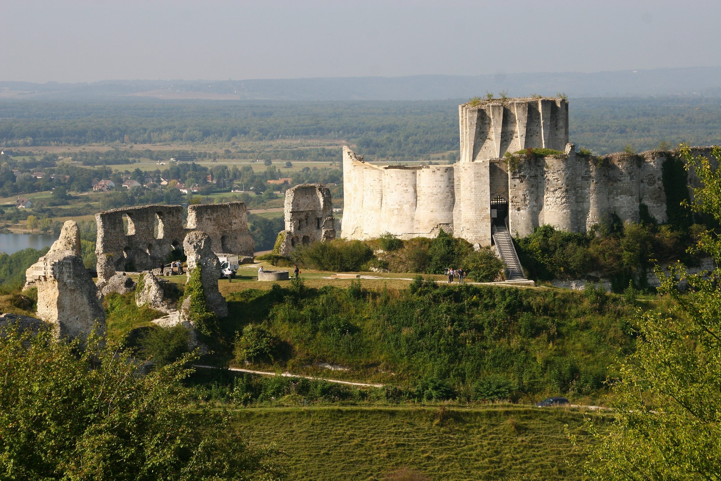 Ruined stone castle stands on a grassy hill, surrounded by lush greenery and overlooking a distant landscape of fields and trees.