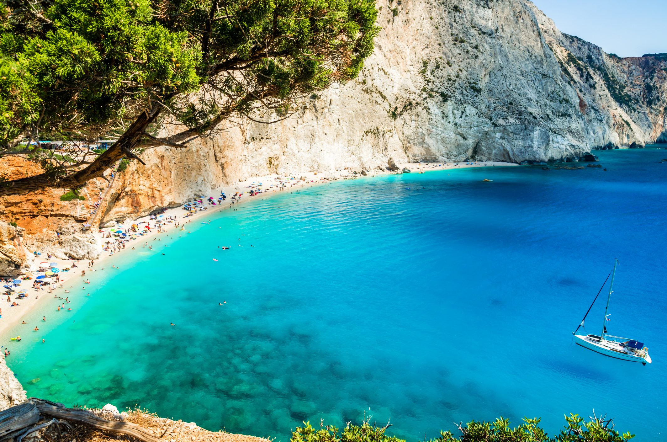 Sailboat floats on clear turquoise water; people relax and swim along vibrant beach. Steep cliffs and overhanging tree frame the coastal scene.