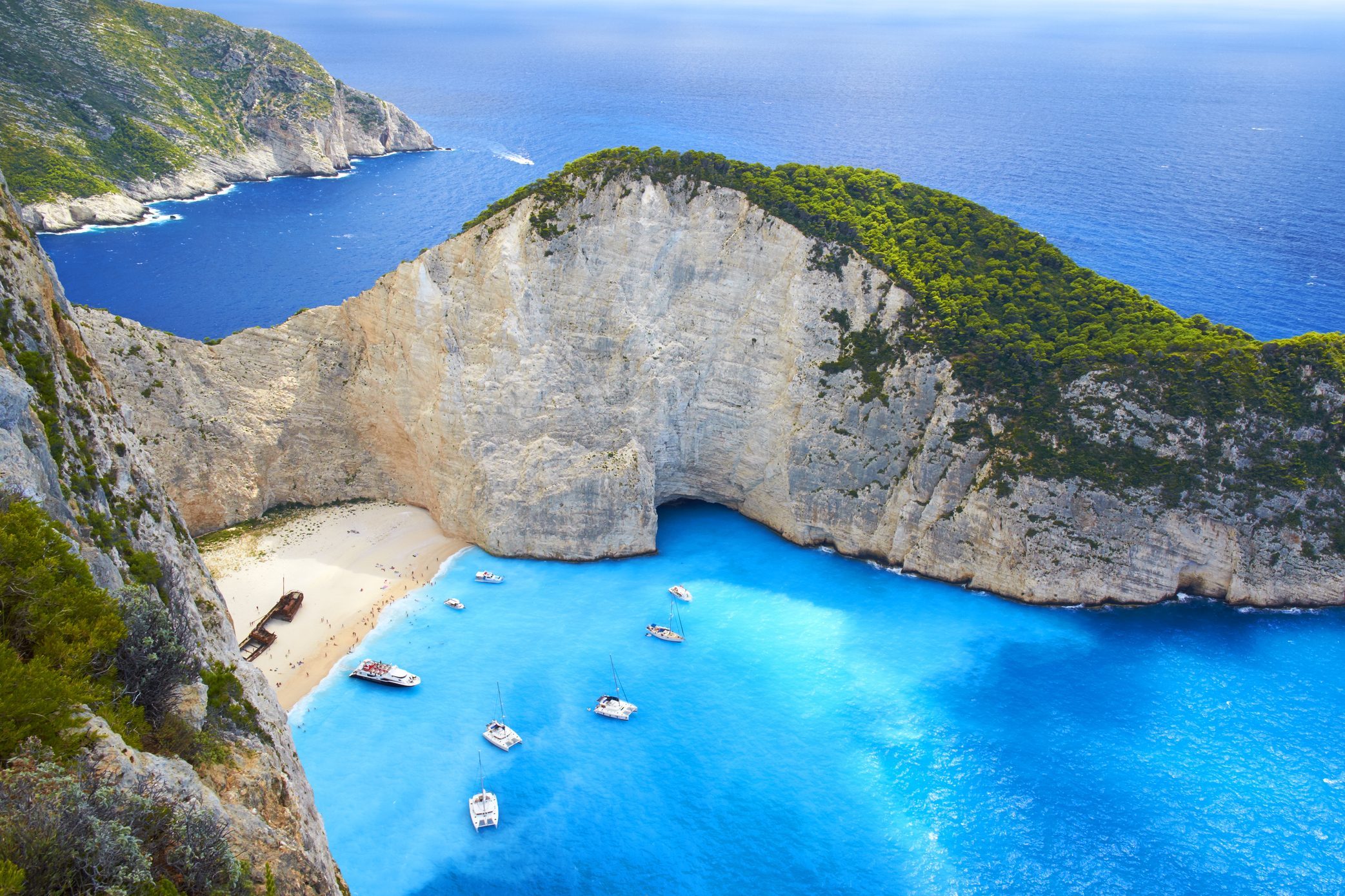 Boats float on vibrant blue waters near a secluded beach, surrounded by steep cliff walls under a clear sky.