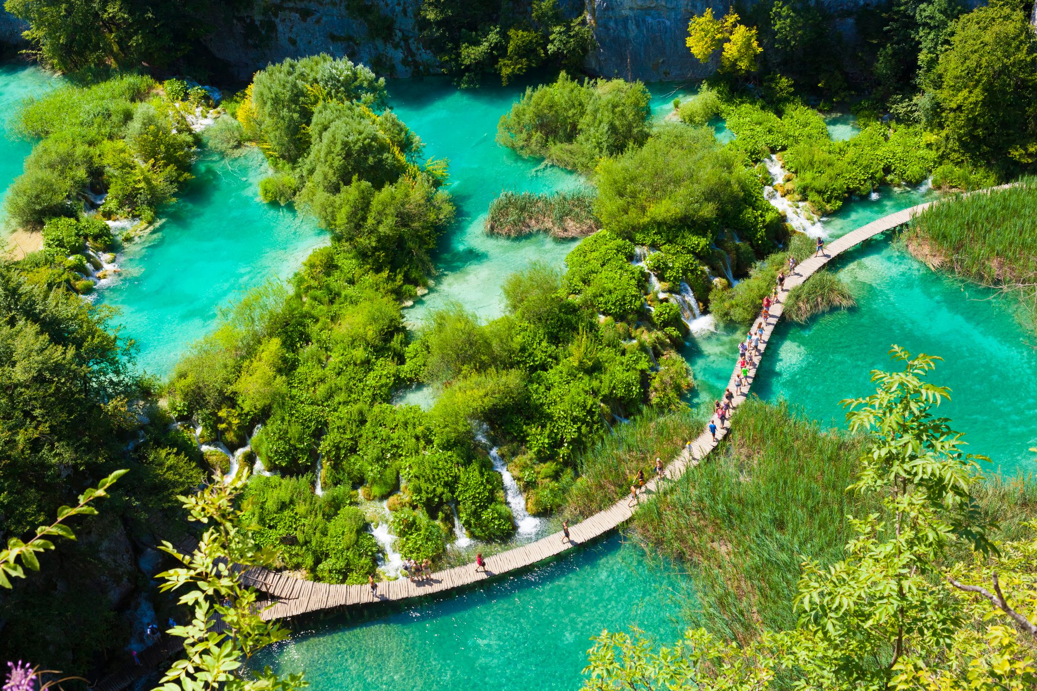 A wooden path curves through vibrant turquoise water with lush green islands. Visitors walk along the path surrounded by waterfalls and dense vegetation.