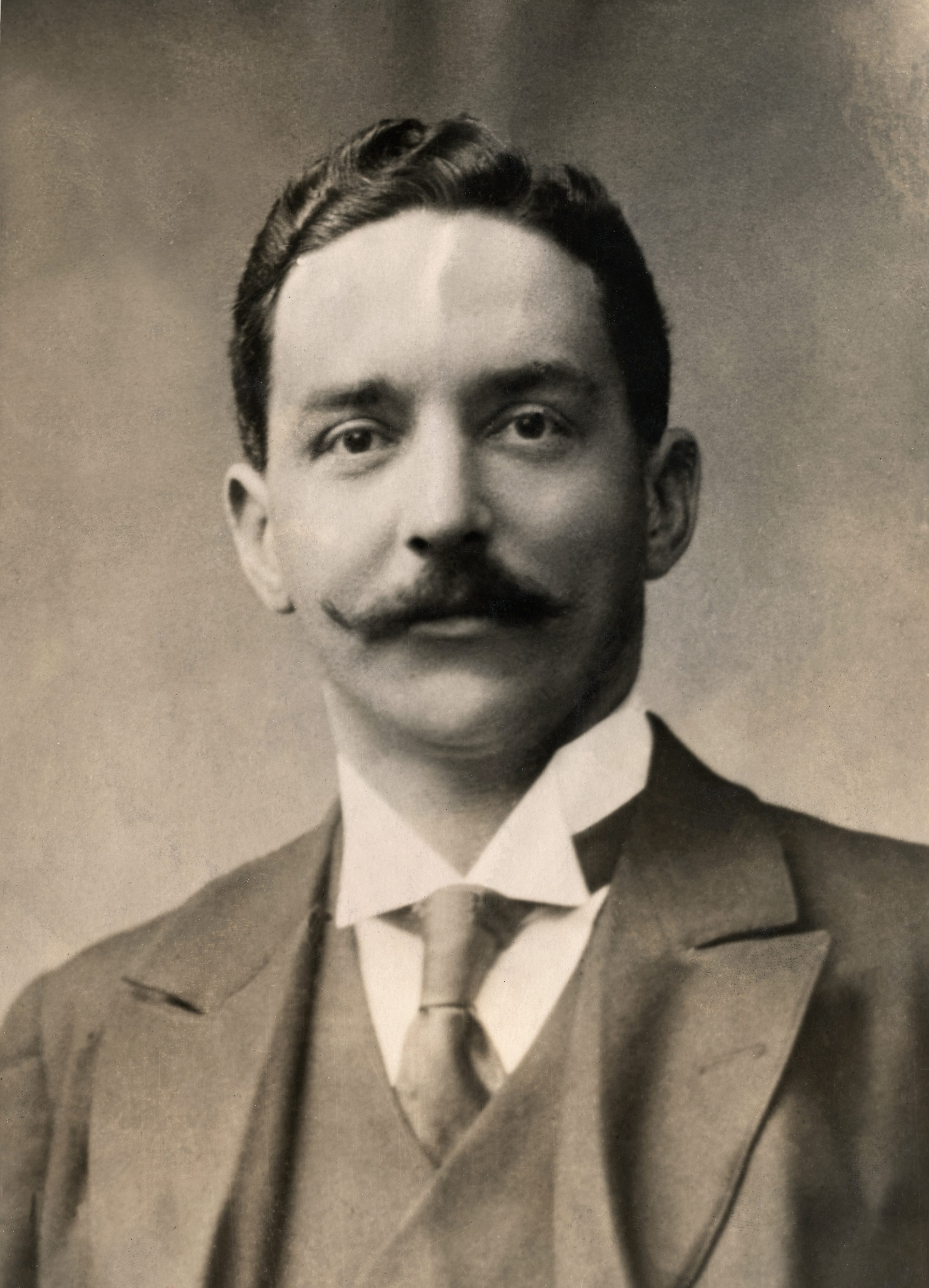 A man with a mustache poses formally, wearing a suit and tie, against a plain background.