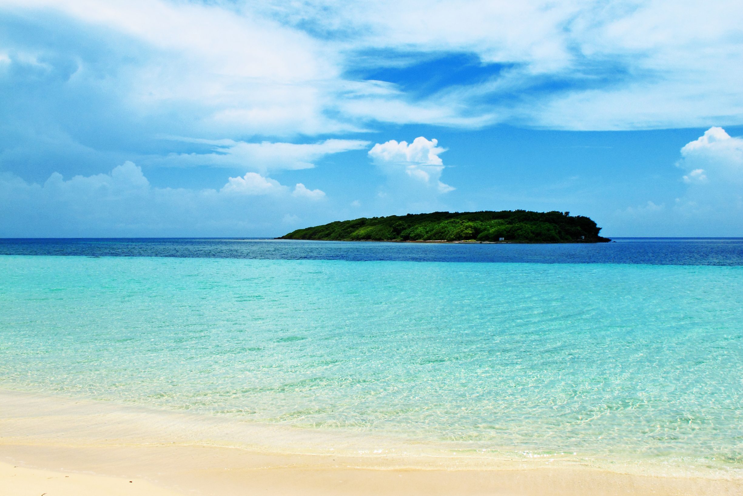 Island sits serenely in the ocean, surrounded by clear turquoise water under a bright blue sky with fluffy white clouds.