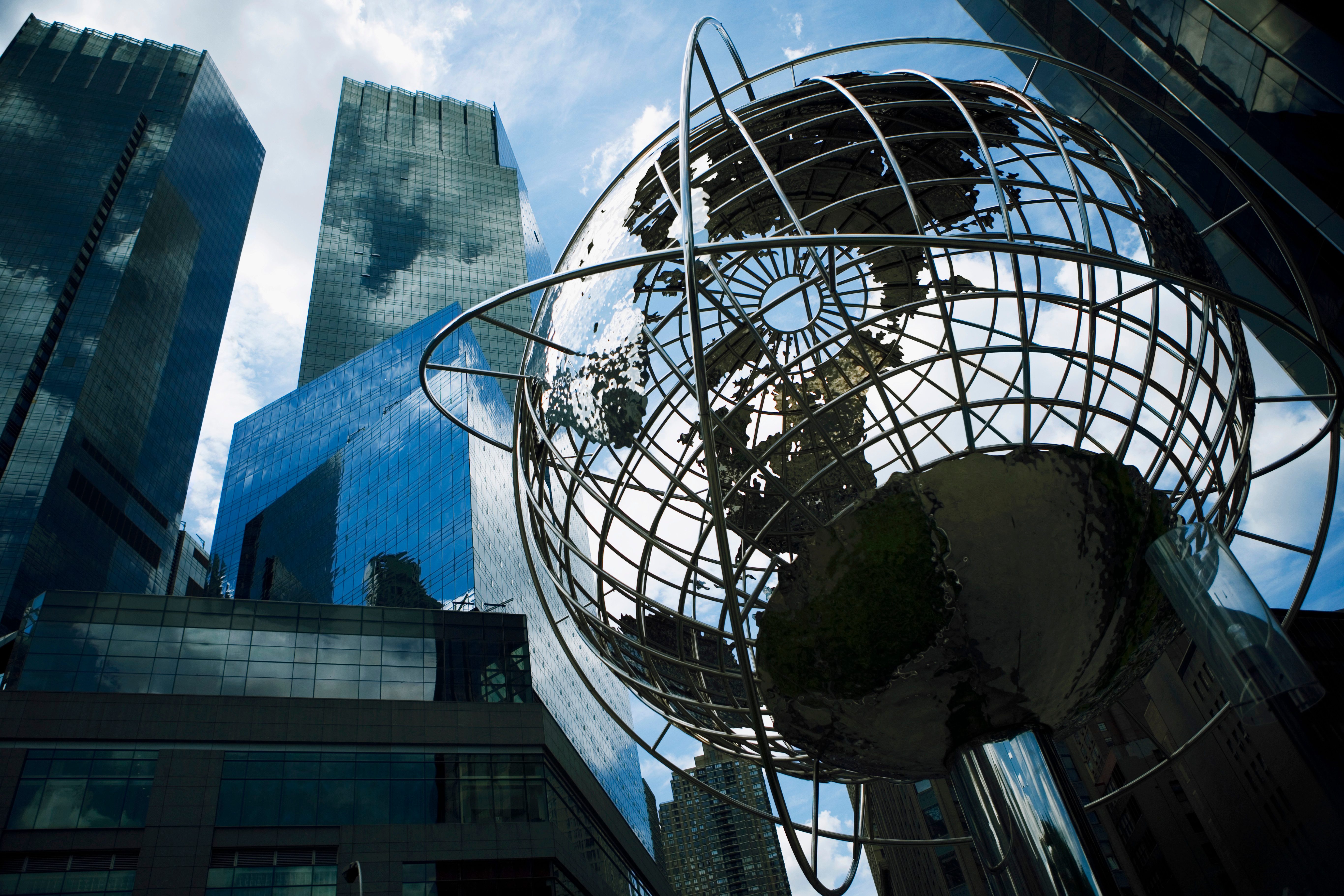Metal globe sculpture reflects sky, surrounded by tall, reflective skyscrapers against a backdrop of blue sky and clouds.