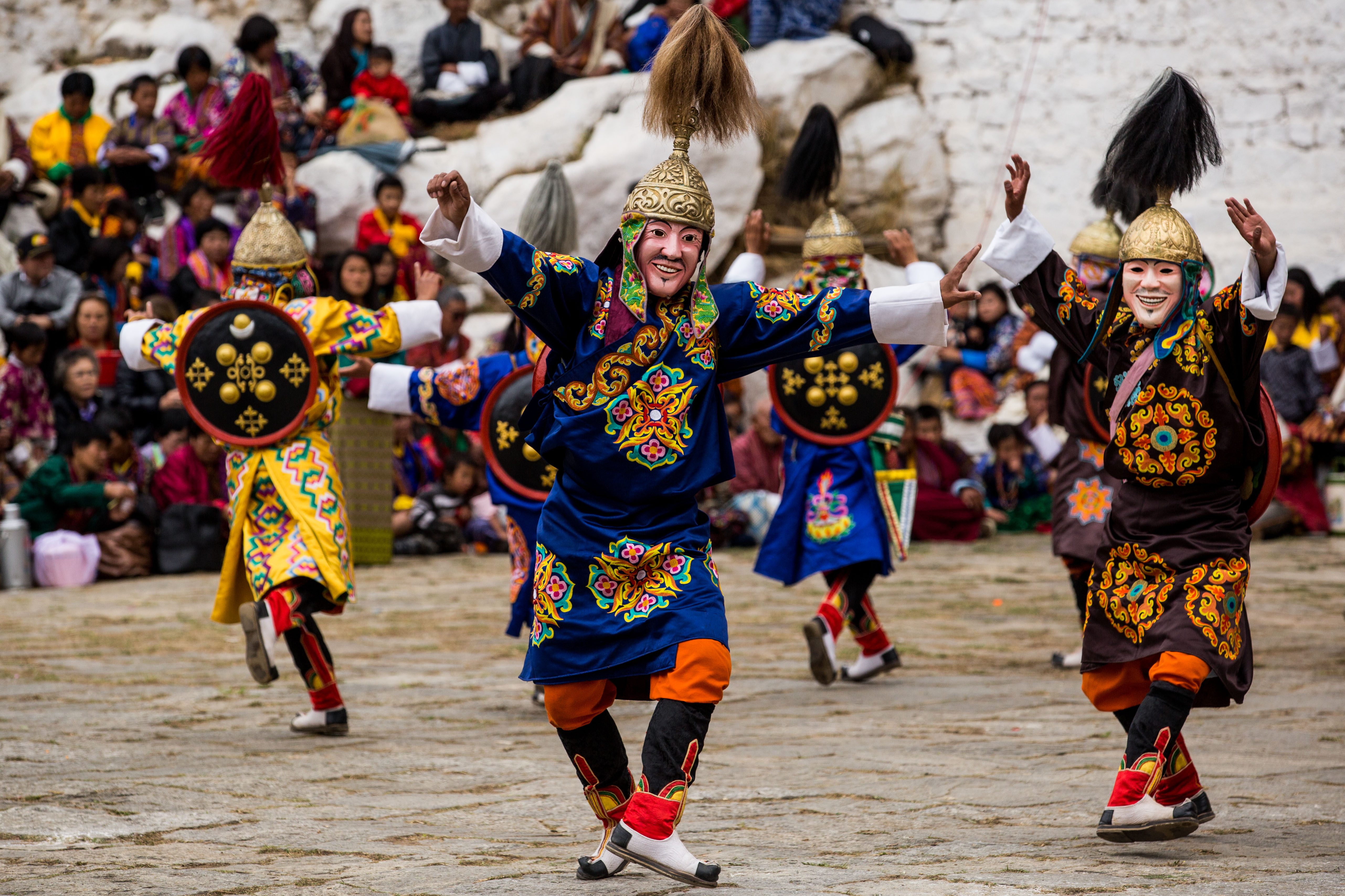 Dancers wear colorful costumes and masks, performing energetically in front of a seated, attentive crowd, set against a stone and mountainous background.