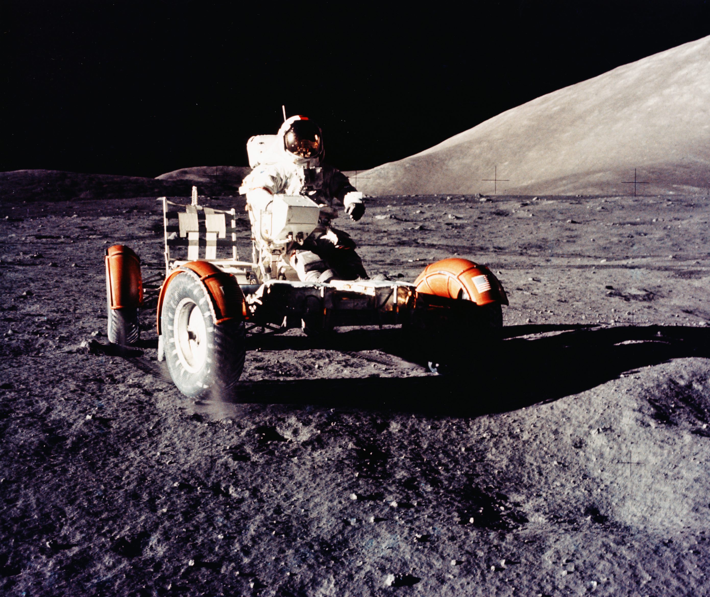 An astronaut drives a moon rover across a grey, dusty lunar surface under a black sky.