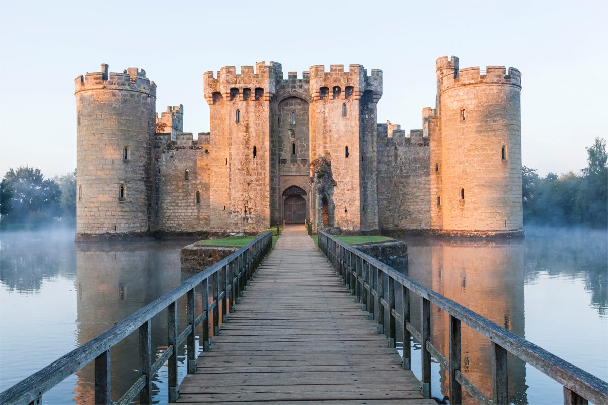 A stone castle stands surrounded by a misty moat, connected by a wooden bridge, reflecting the soft morning light.