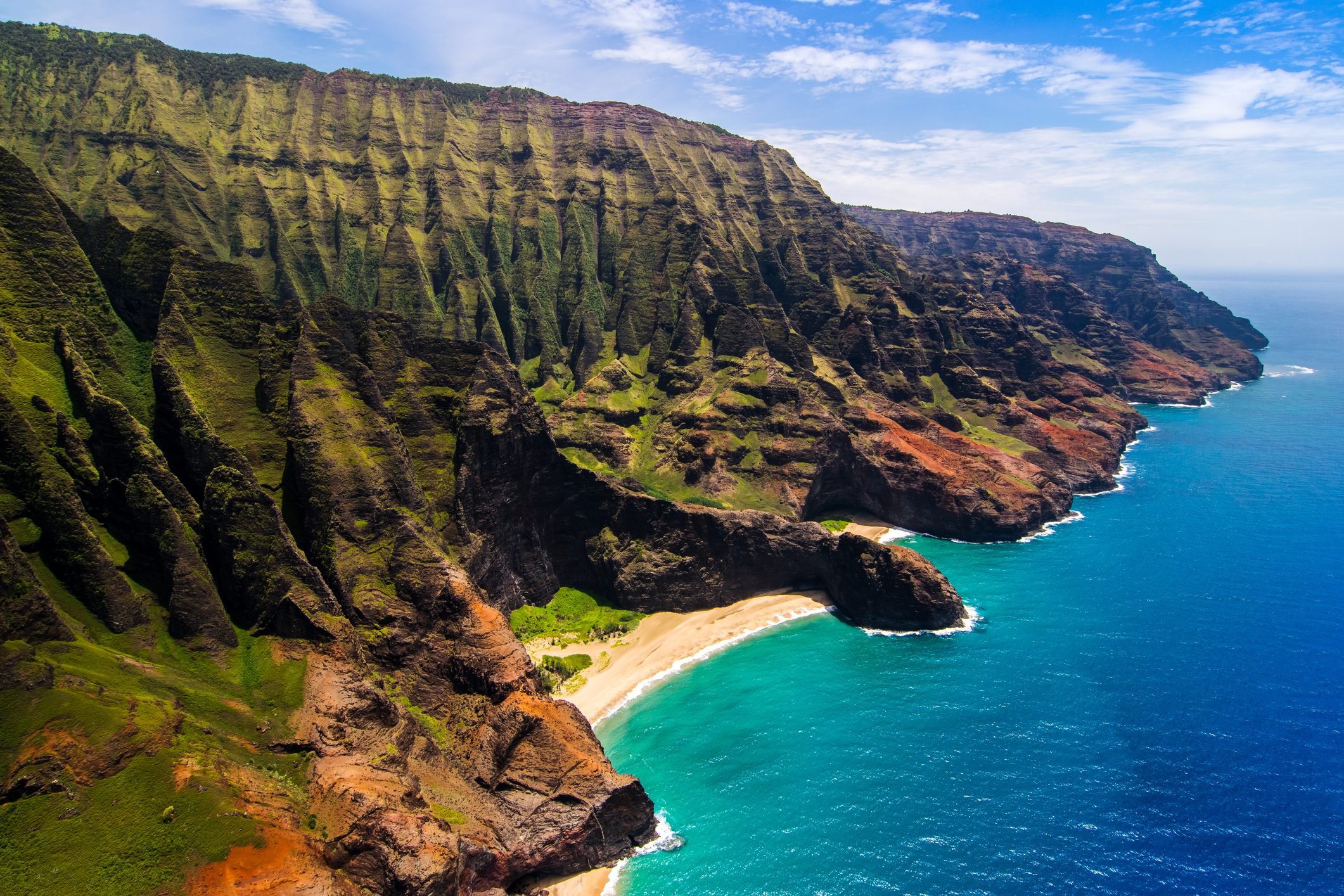 Rugged coastal cliffs rise above turquoise ocean waters, bordered by a sandy beach under a bright blue sky.