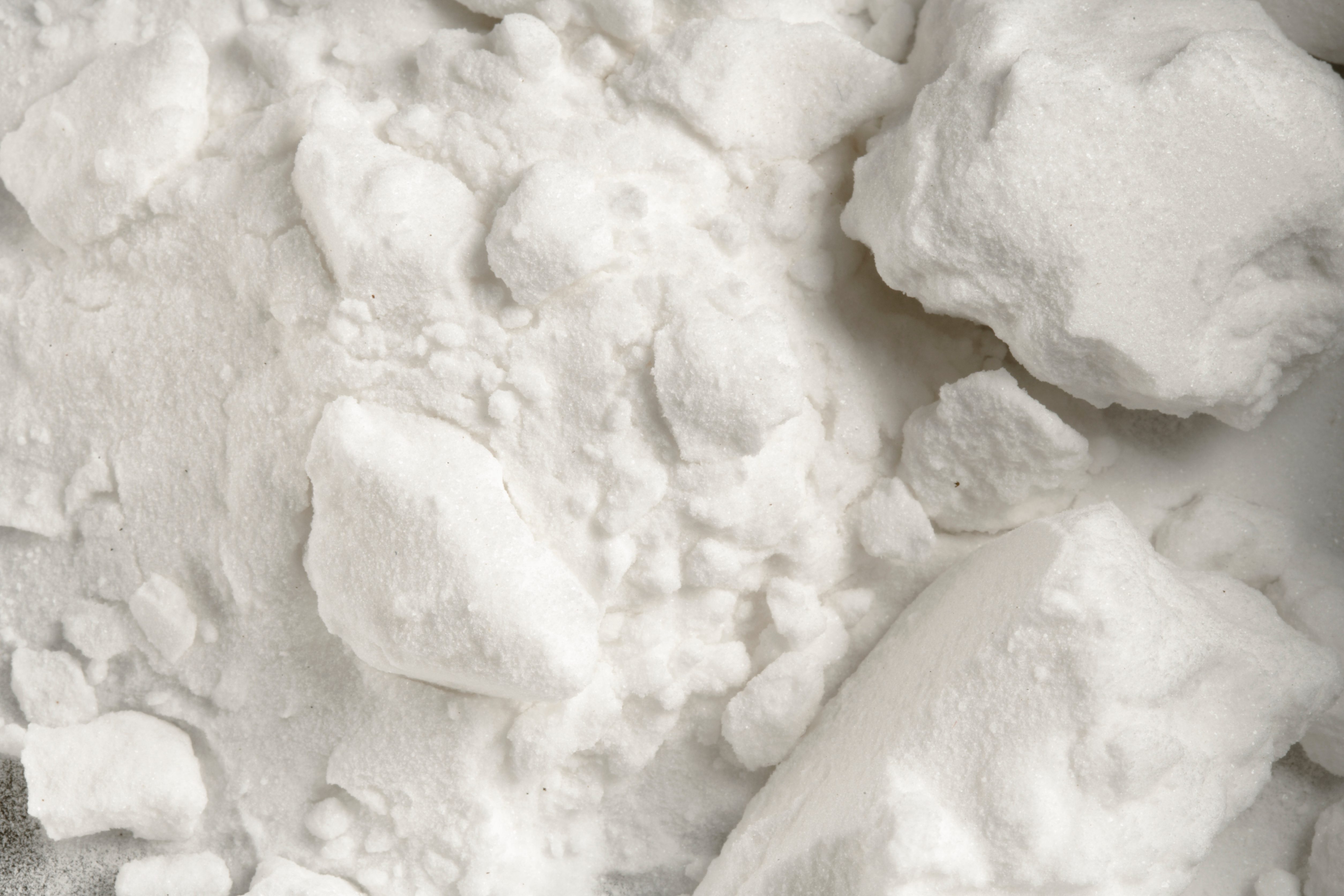 White crystalline powder resting in clusters on a flat surface, appearing coarse in texture.
