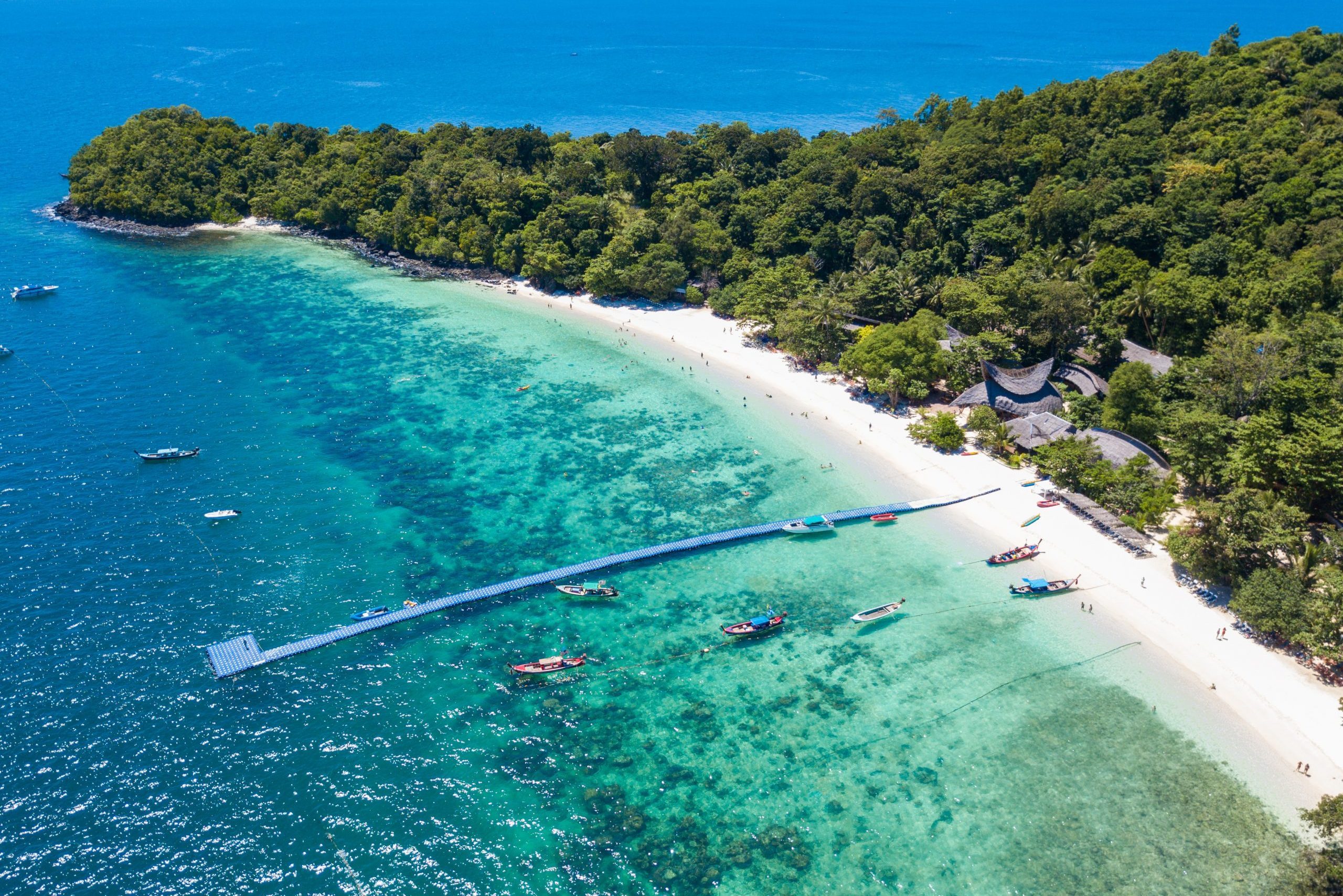 Boats float near a long, floating pier along a tropical beach with lush greenery in the background, surrounded by clear turquoise water.