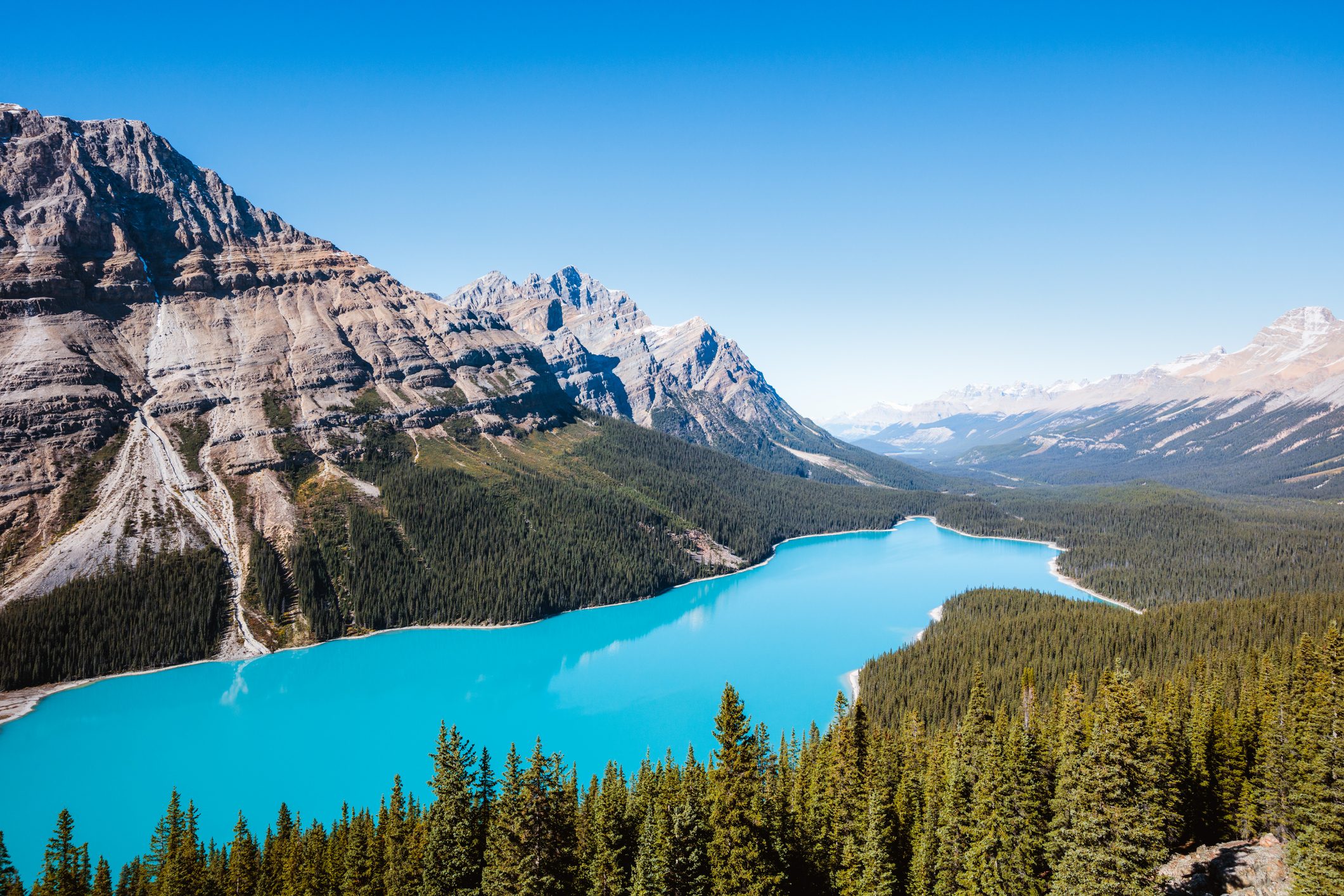 A turquoise lake rests amid dense forests, bordered by jagged mountains under a clear blue sky.