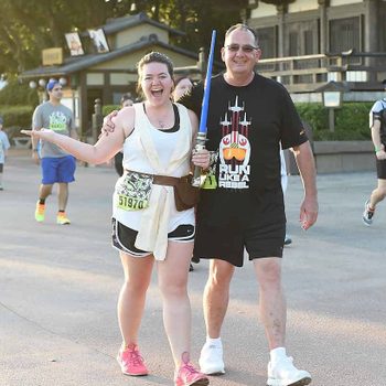 Participants smile while holding a lightsaber, walking in a race event near a traditional Asian building. Shirts read "RUN LIKE A REBEL."