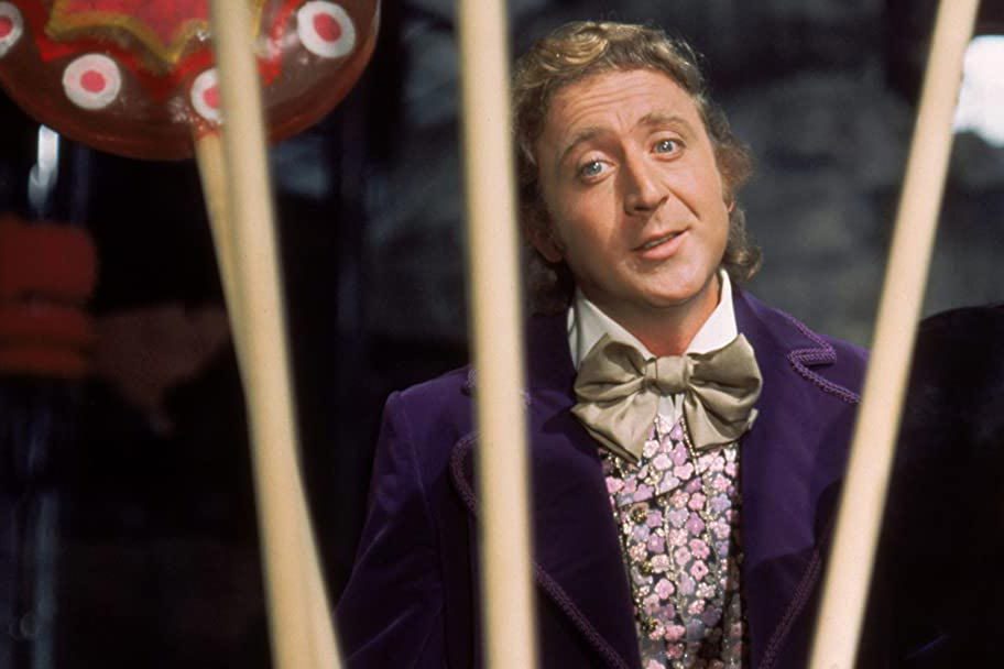 A man wearing a purple coat and bow tie stands behind oversized candies in a whimsical, colorful setting.