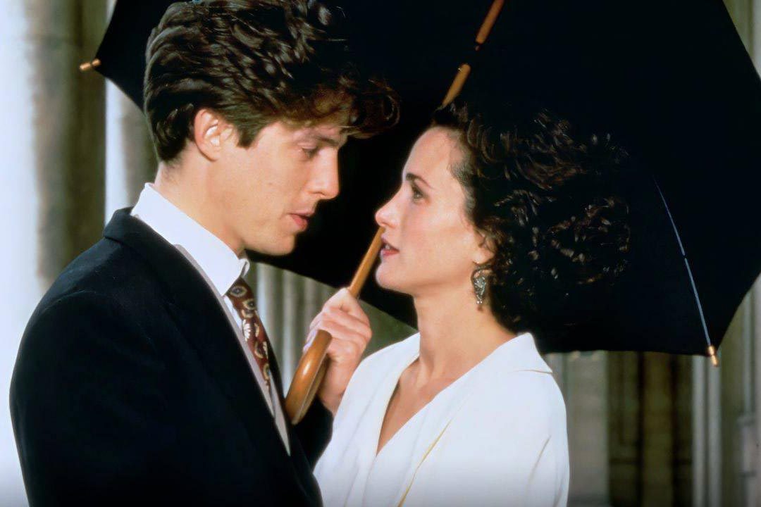 Two people stand closely, holding an umbrella, indoors. One wears a suit; the other, a white outfit. They seem engaged in conversation.