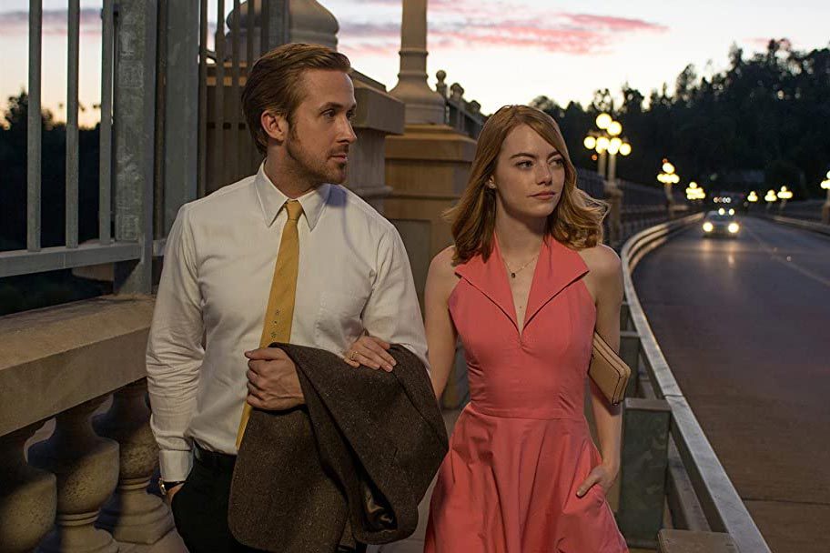 A man and woman walk on a bridge at dusk, illuminated by streetlights, with cars approaching in the background.