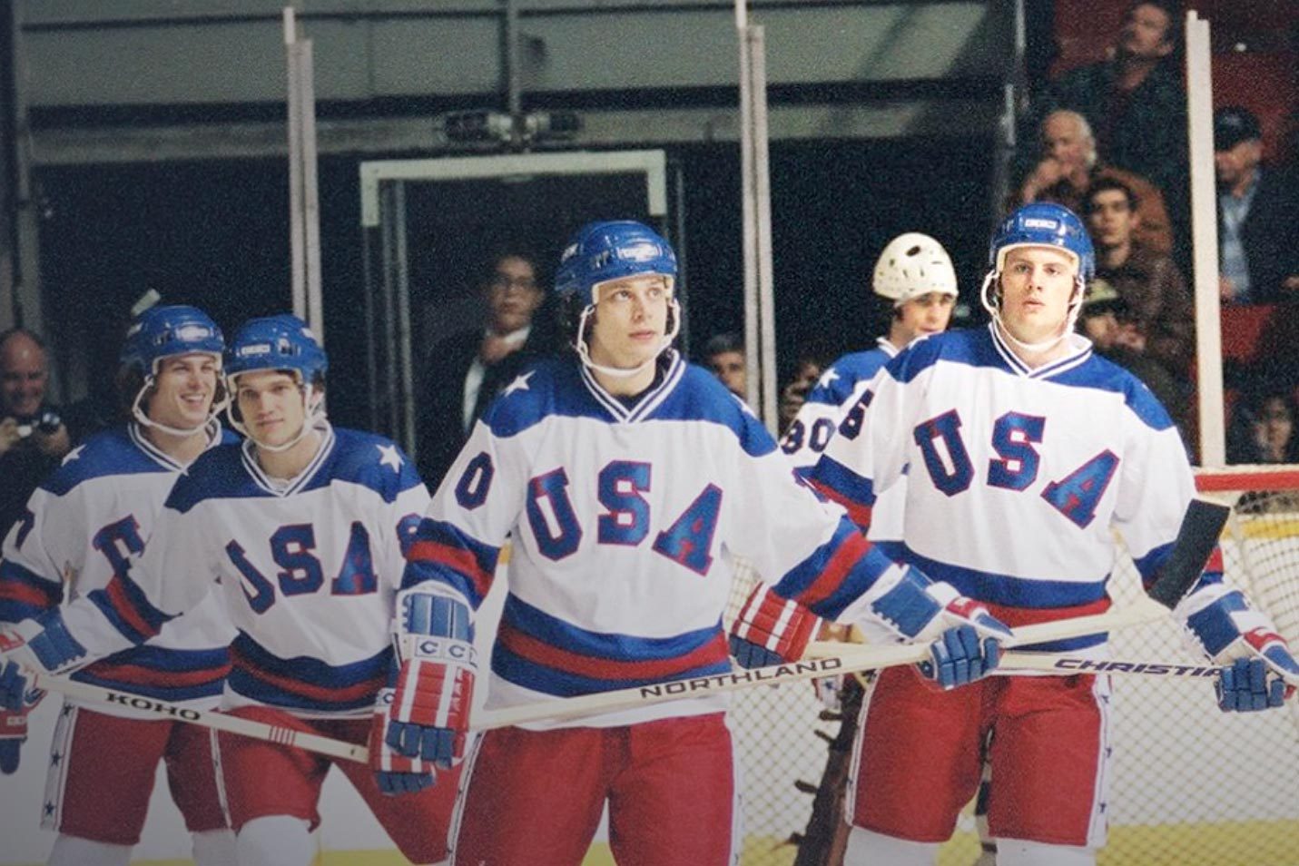 Hockey players walk on ice rink wearing white jerseys with "USA," blue helmets, and gloves. Spectators visible in the background.