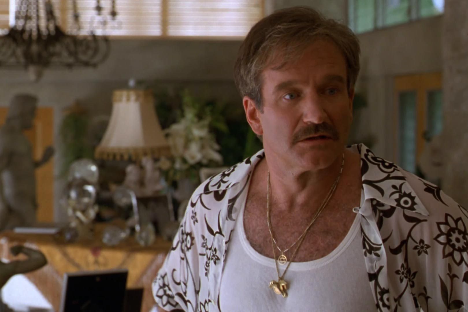 Man stands in ornate room, wearing a floral shirt and necklace, with lamps, plants, and statues in the background.