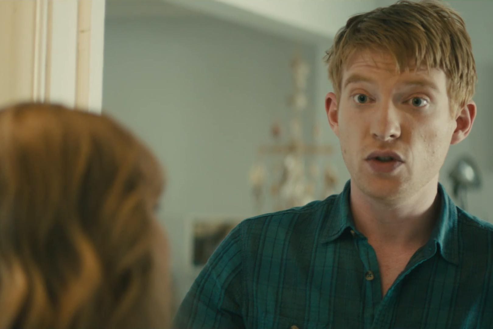 A man in a plaid shirt speaks to a woman in an indoor setting with muted colors.