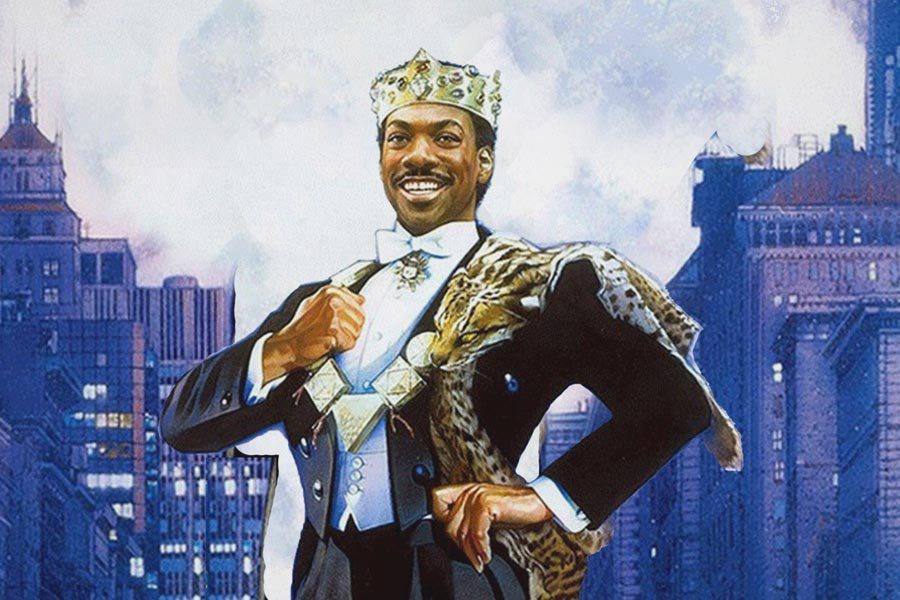 A man wears a crown and regal outfit, smiling confidently with city skyscrapers in the background.