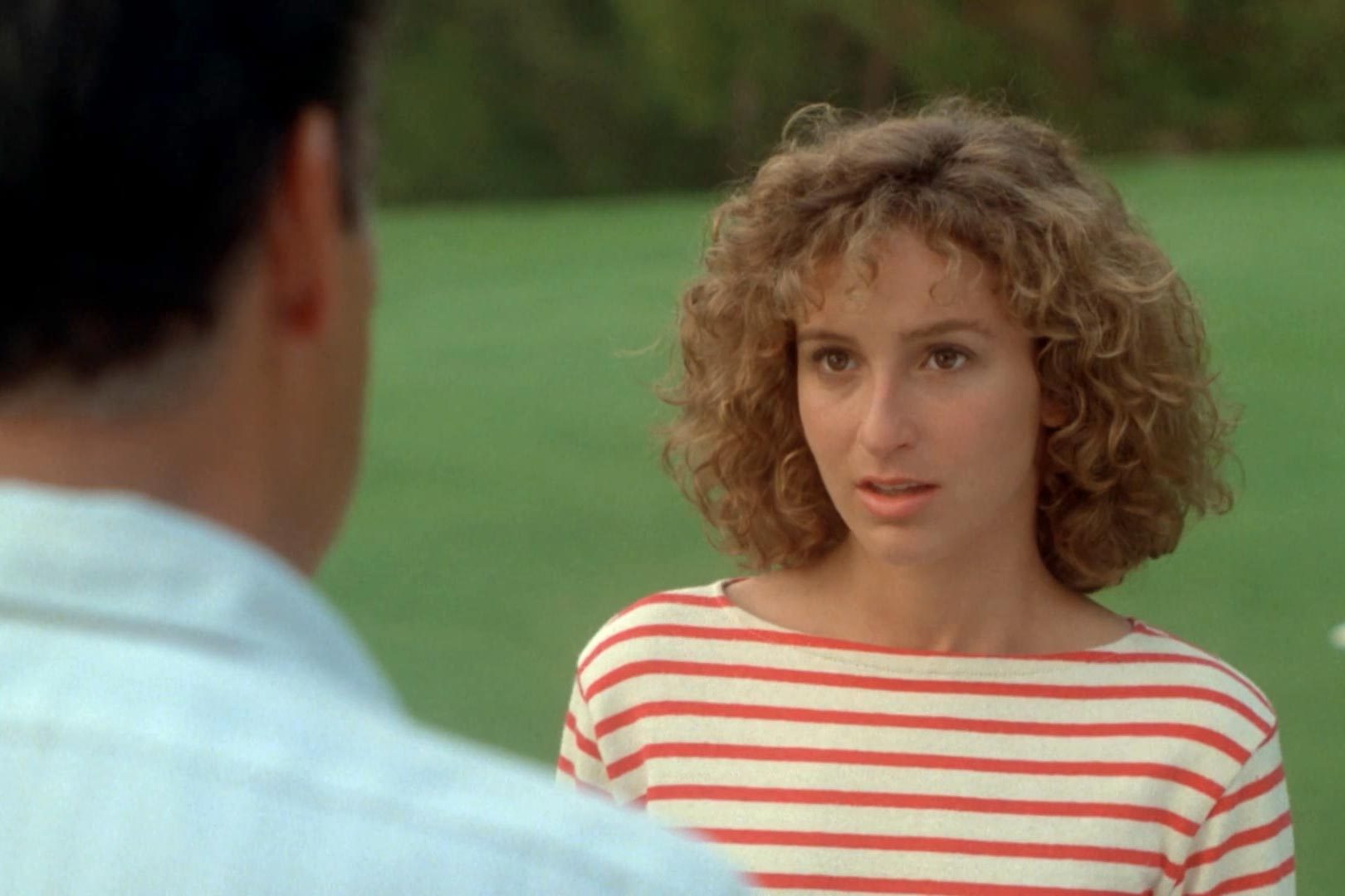 Woman stands outdoors, wearing a striped shirt, looking at someone off-camera, with grass and trees in the background.