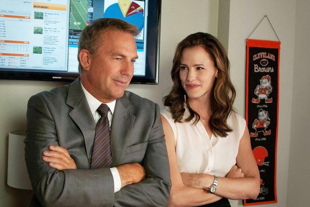 Two people stand smiling, arms crossed, in an office with sports statistics on monitors and a Cleveland Browns banner on the wall.