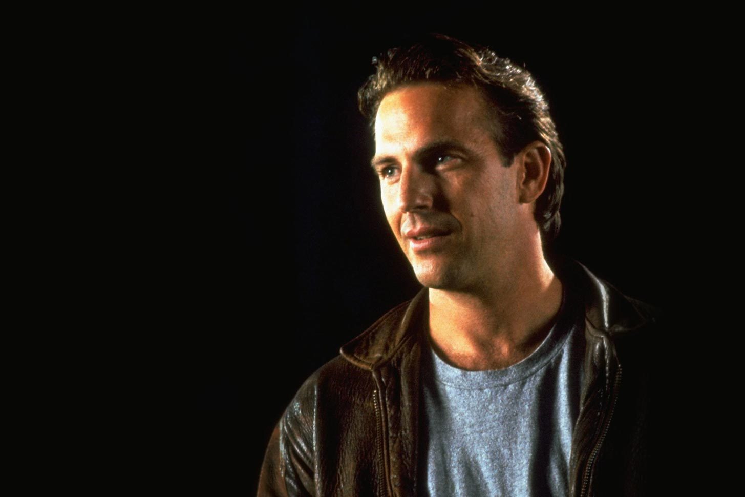 A man stands with a leather jacket in a dark environment, looking to the side, visible from the chest up.