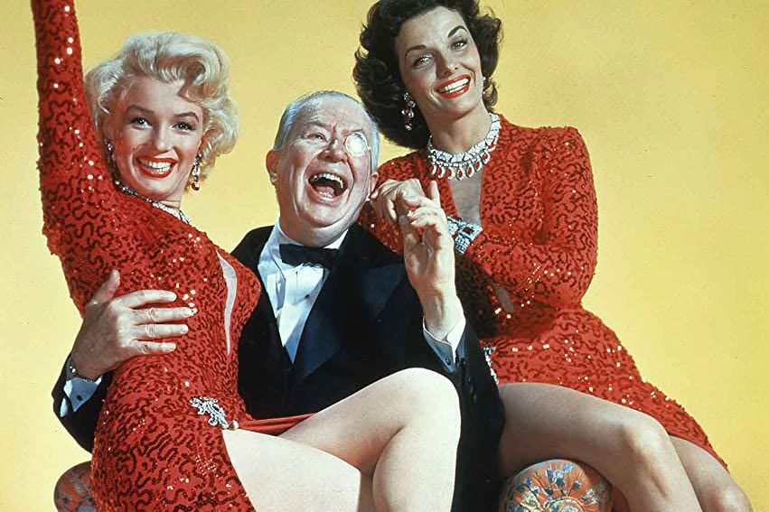 Three people pose cheerfully; two women in red sequin dresses sit beside a man in a suit, laughing against a plain yellow background.