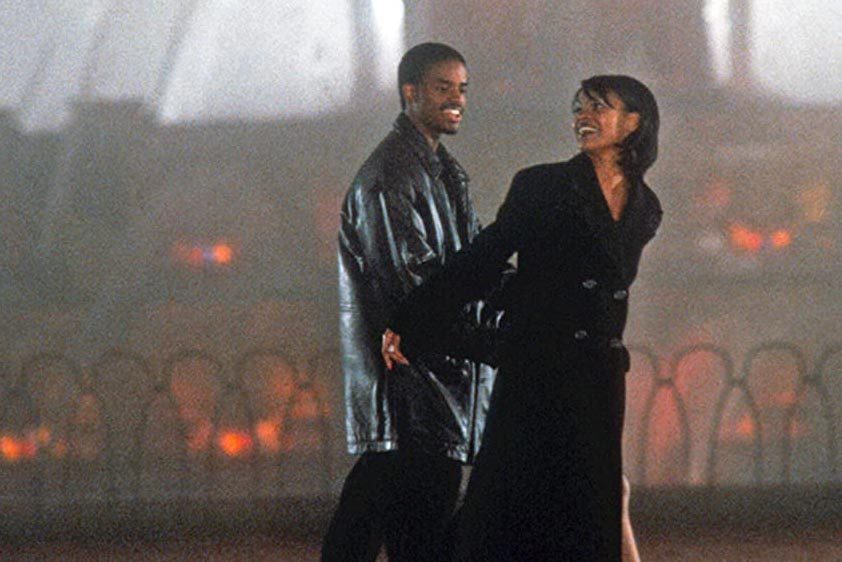 Two people wearing coats walk together, smiling, in a foggy, softly lit outdoor setting with blurred lights in the background.