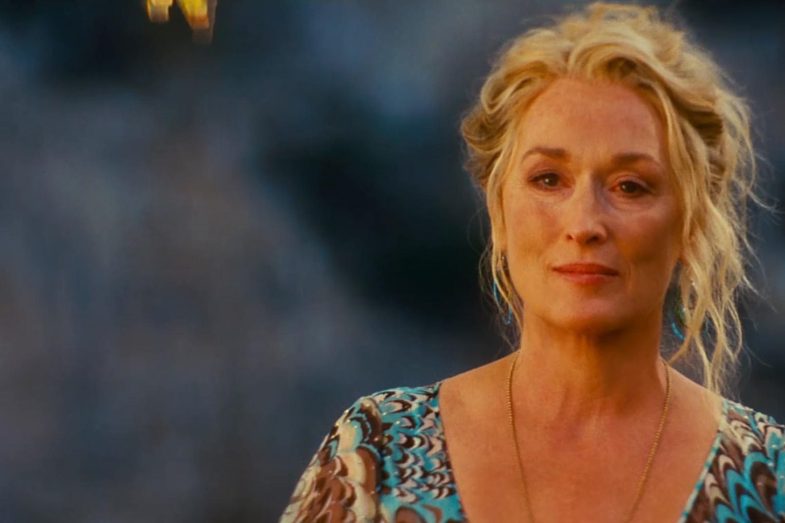 Woman stands serenely, wearing a patterned top, against a blurred outdoor background, creating a calm and introspective atmosphere.