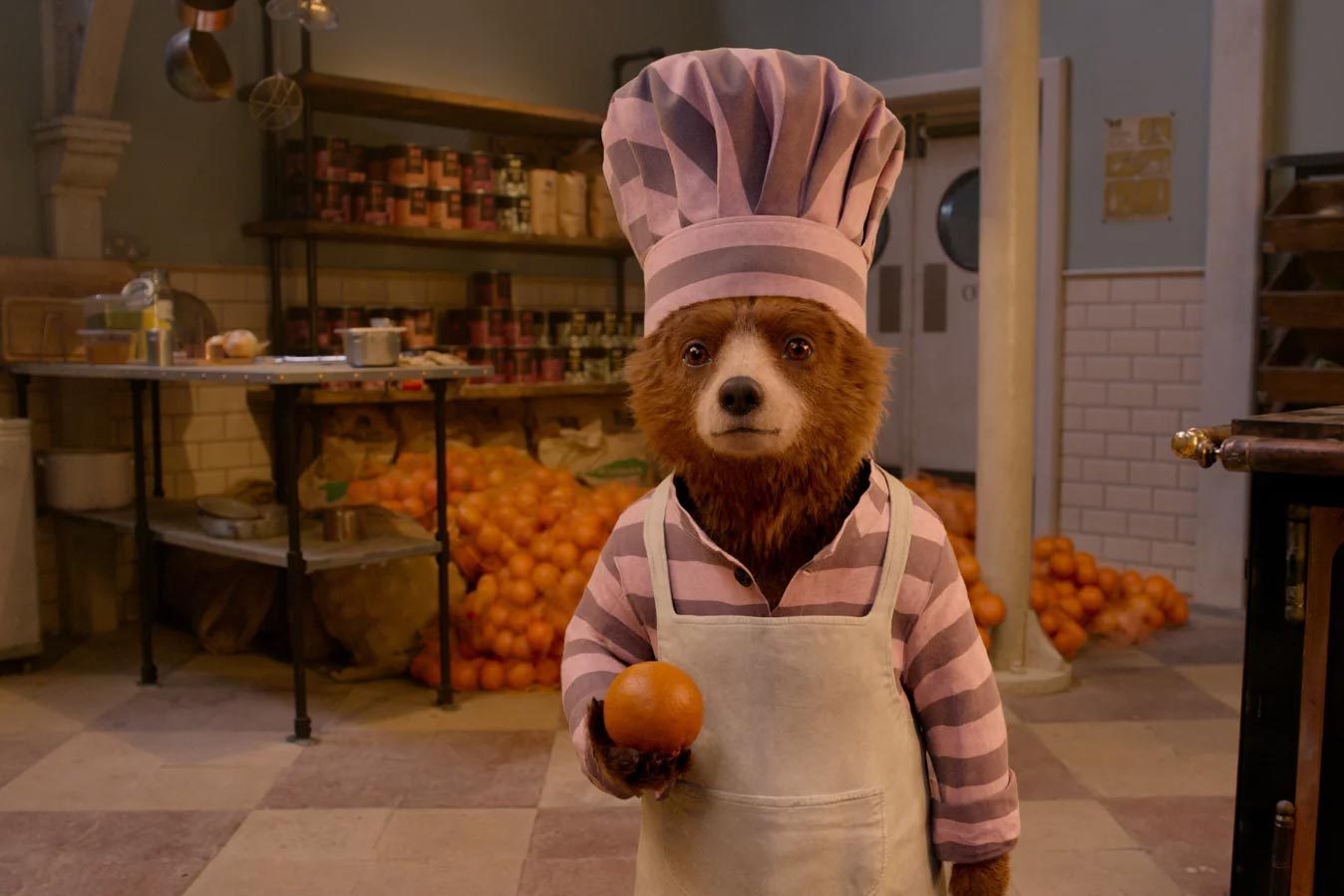 A bear wearing a chefâ€™s hat holds an orange in a kitchen filled with stacked pots, pans, and piles of oranges.