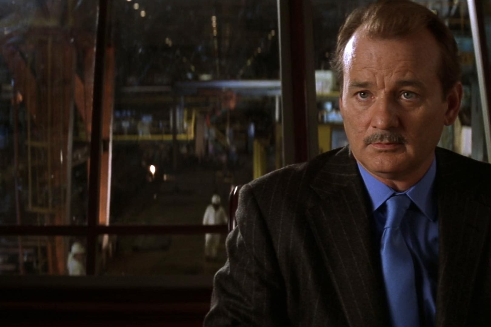 A man in a suit observes quietly in an industrial setting, with workers and machinery visible in the background.