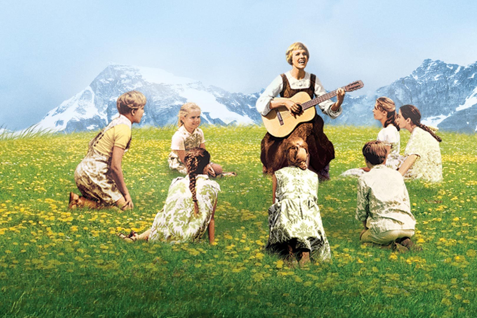 A woman plays guitar surrounded by children sitting on a grassy field with yellow flowers, with snow-capped mountains in the background.