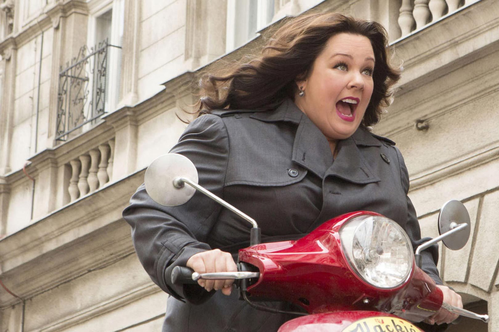 A woman rides a red scooter with excitement on her face, passing alongside a stone building with ornate railings.