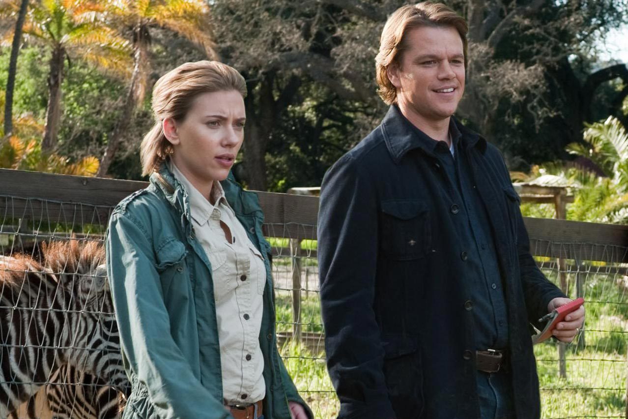 Two people stand by a fence with a zebra, surrounded by lush greenery and trees in a zoo-like environment.