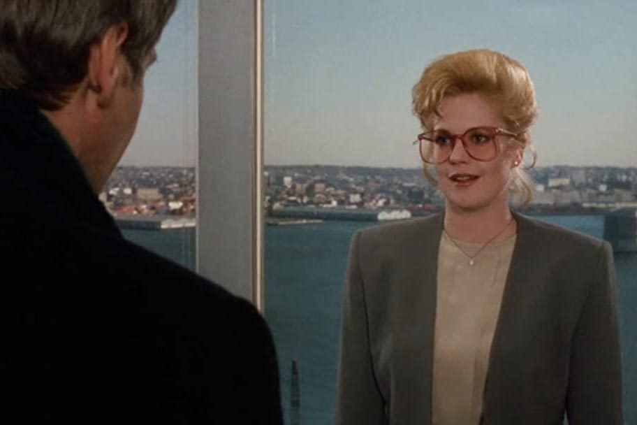 A woman with large glasses talks to a man, set against a city skyline view through a large window.