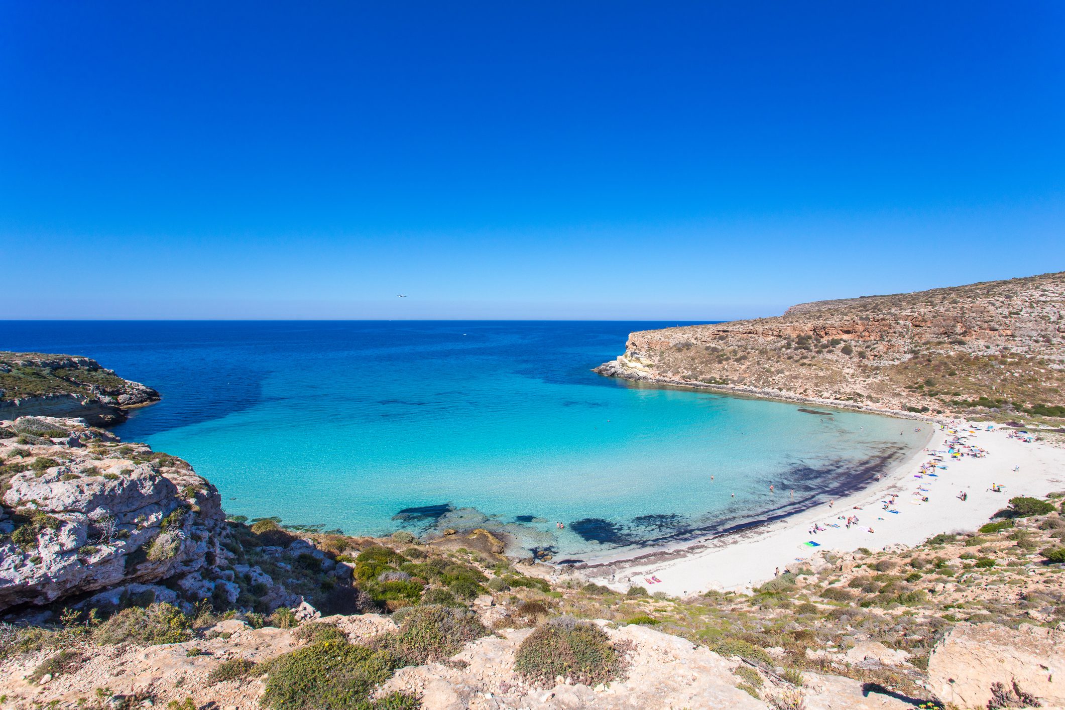 Turquoise water rests calmly against a sandy beach, surrounded by rugged cliffs under a clear blue sky. Beachgoers relax along the shoreline.