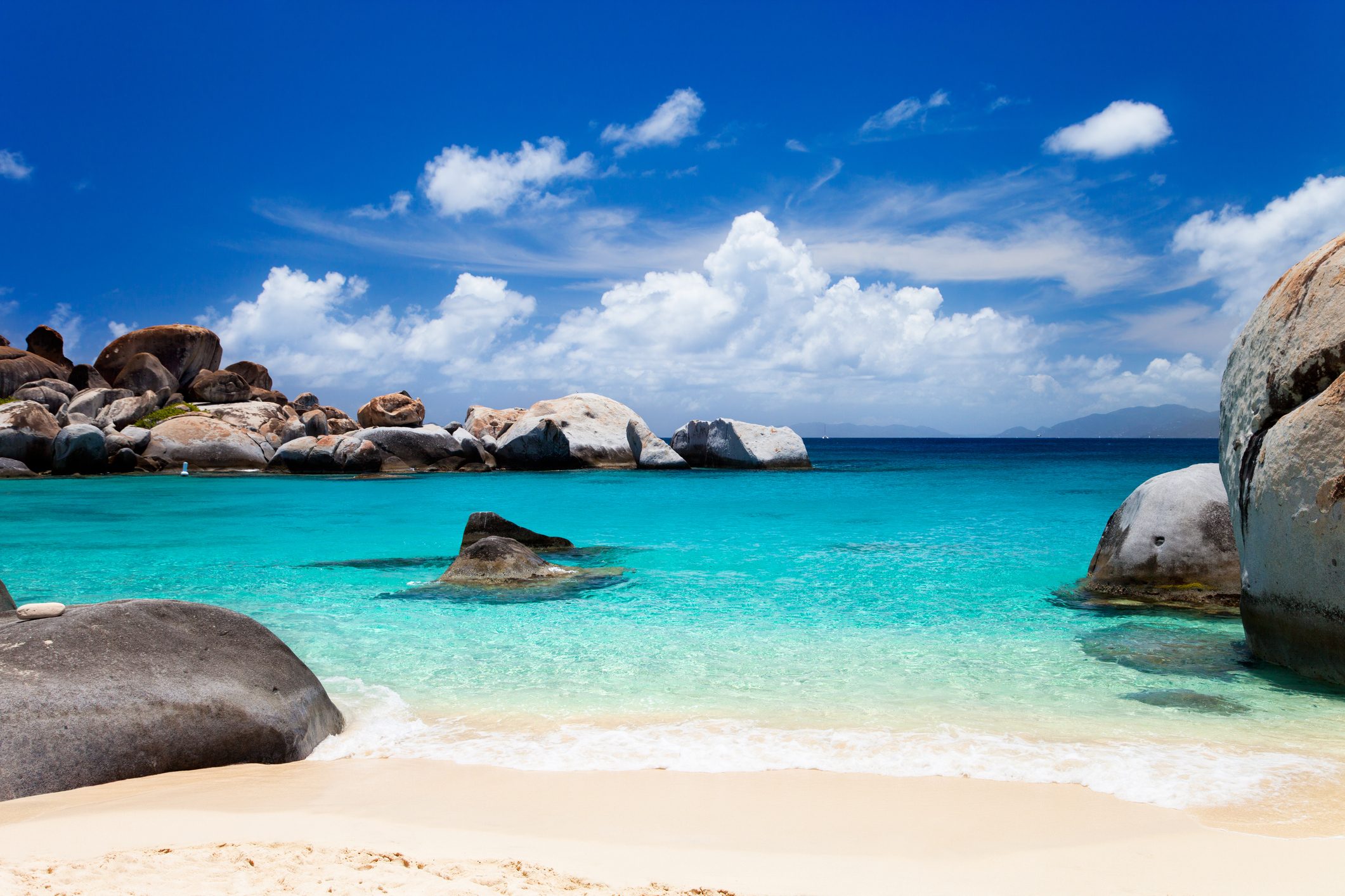 Boulders rest on a sandy beach with turquoise water, under a blue sky scattered with fluffy clouds.