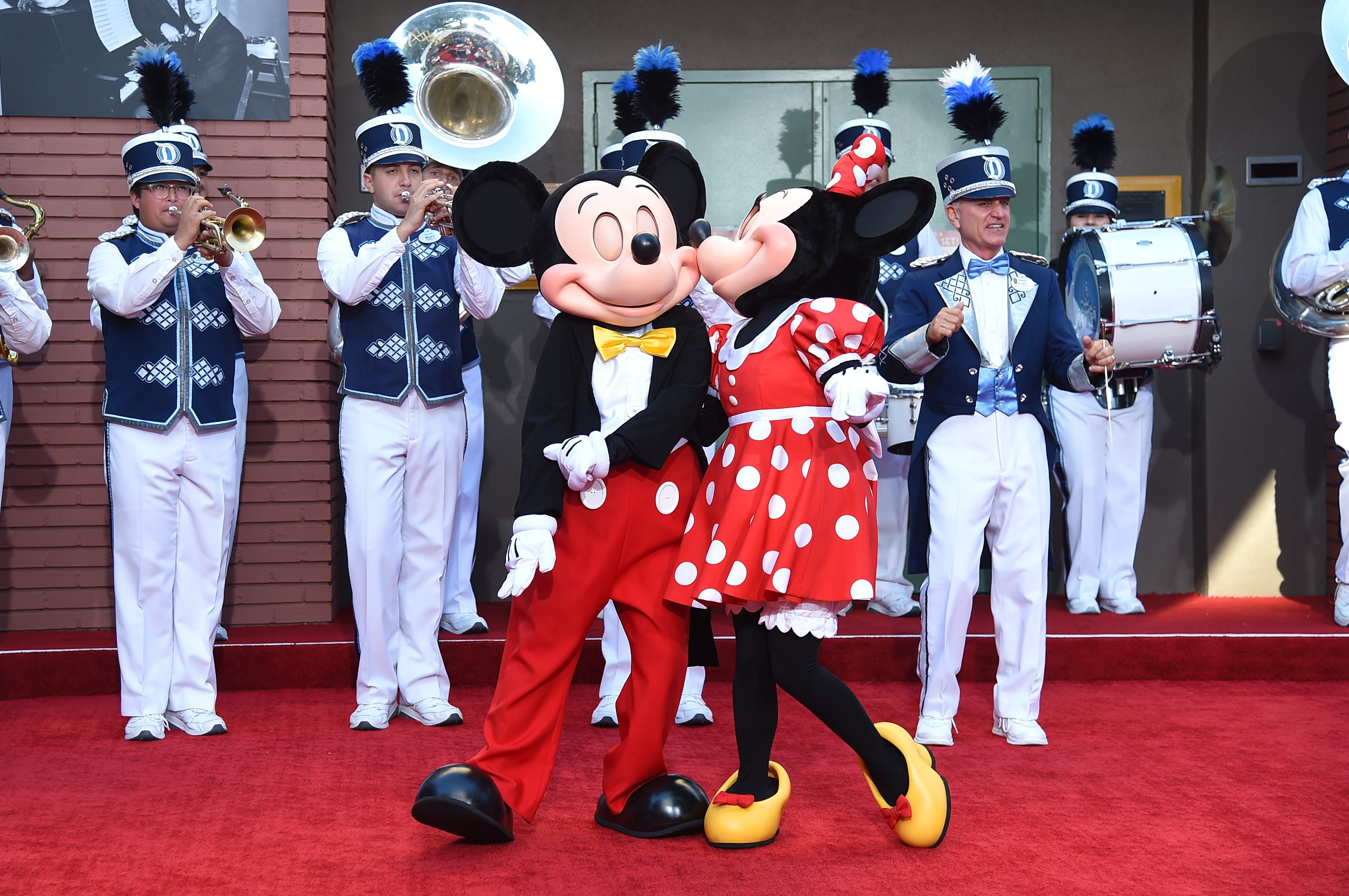 Two mascots affectionately pose on a red carpet, surrounded by a marching band playing instruments.