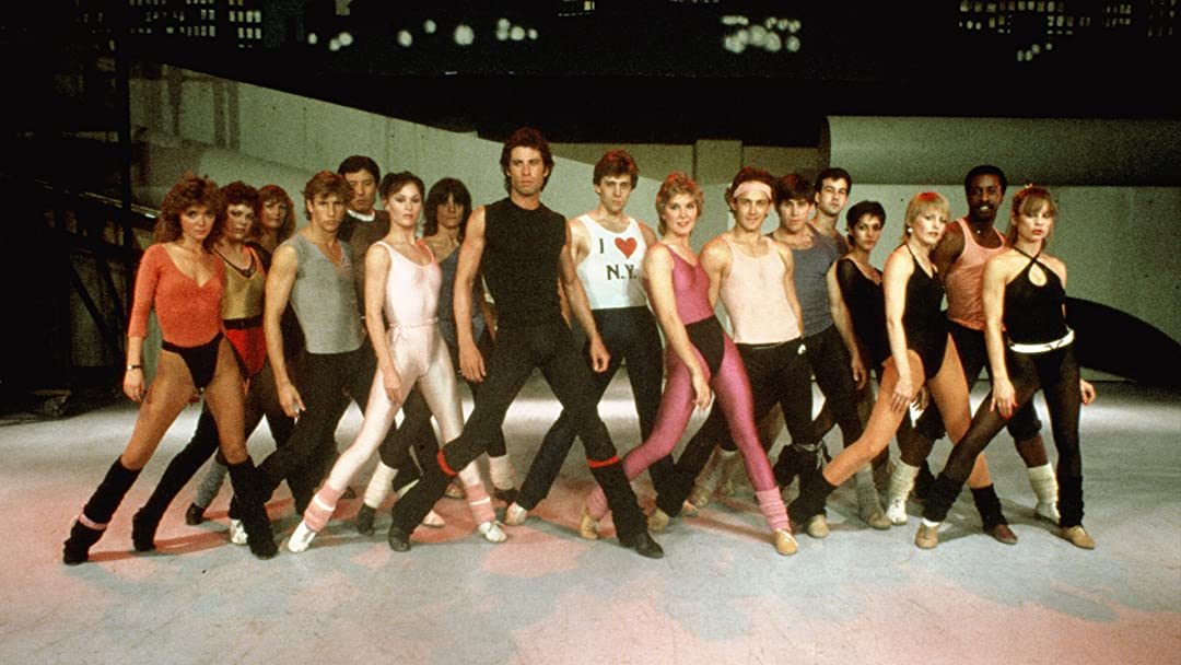 Dancers stand posing in a line wearing colorful leotards, set against a backdrop resembling a cityscape. One dancer's shirt reads "I â™¥ N.Y."